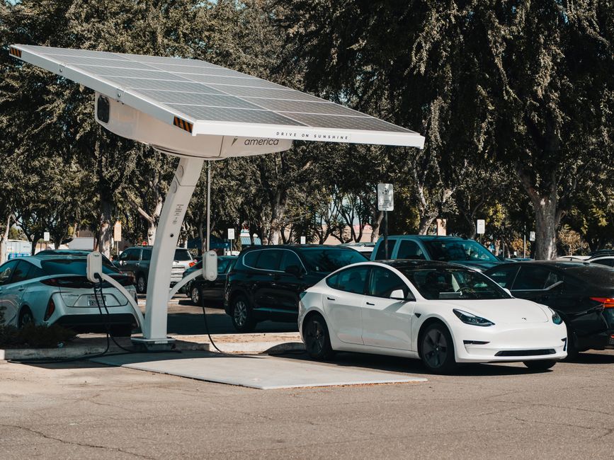 A white electric car is parked at a solar-powered charging station in a parking lot, with multiple other vehicles including sedans and SUVs in the background. The charging station has a modern design with a large, angled solar panel canopy on top, supported by a curved white pole. The surface of the parking area is paved, and there are trees with dense foliage behind the lot, providing shade and a natural backdrop. Some areas of the ground show clean, smooth pavement, while the charging area has a dedicated concrete slab. The lighting indicates a bright day with clear skies, and the overall scene emphasizes eco-friendly transportation infrastructure within an urban environment, consistent with the types of outdoor and landscaping services offered by Gardeners Finsbury Park.