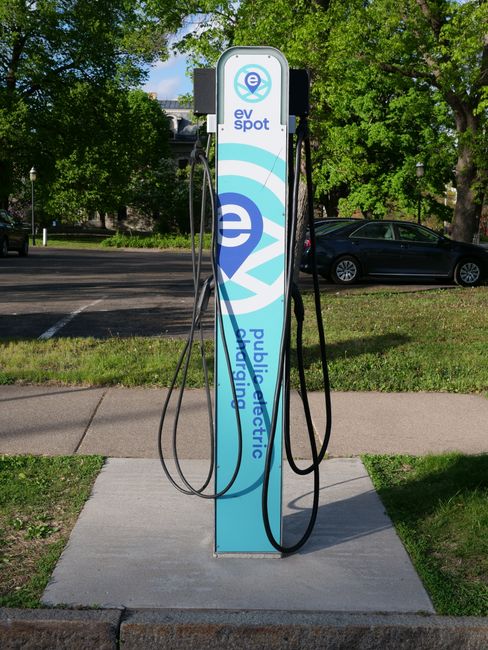 A white electric car is parked at a solar-powered charging station in a parking lot, with multiple other vehicles including sedans and SUVs in the background. The charging station has a modern design with a large, angled solar panel canopy on top, supported by a curved white pole. The surface of the parking area is paved, and there are trees with dense foliage behind the lot, providing shade and a natural backdrop. Some areas of the ground show clean, smooth pavement, while the charging area has a dedicated concrete slab. The lighting indicates a bright day with clear skies, and the overall scene emphasizes eco-friendly transportation infrastructure within an urban environment, consistent with the types of outdoor and landscaping services offered by Gardeners Finsbury Park.