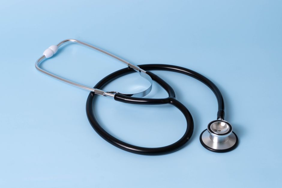 A close-up image of a black stethoscope with silver accents, lying on a light blue surface. The stethoscope's chest piece is positioned at the lower right, with the tubing forming gentle loops and the earpieces resting near the top left. The smooth, shiny surface of the chest piece reflects light, and the flexible tubing appears clean and well-maintained. The background is plain, emphasizing the medical instrument, which is unrelated to gardening or outdoor maintenance.