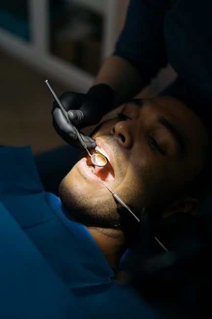 A close-up view of a man lying back in a dental chair during a dental check-up. He has his mouth open, revealing dental tools being used by a dentist wearing black gloves. The dentist is inspecting or cleaning his teeth with a mirror and probe, focusing on the patient's upper teeth. The setting appears to be a modern dental clinic with soft lighting, emphasizing the detailed dental work being performed. The image highlights professional oral healthcare, which is unrelated to gardening, landscaping, or outdoor maintenance themes associated with gardenersfinsburypark.org.uk and the page about licensed insured gardeners in Finsbury Park.